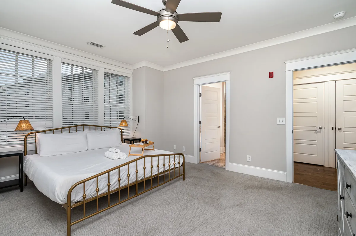 A serene bedroom featuring a bed with a golden frame, flanked by two bedside lamps and complemented by light wooden furniture and open doorways. Sunlight filters gently through the room, creating an inviting, restful atmosphere ideal for unwinding.