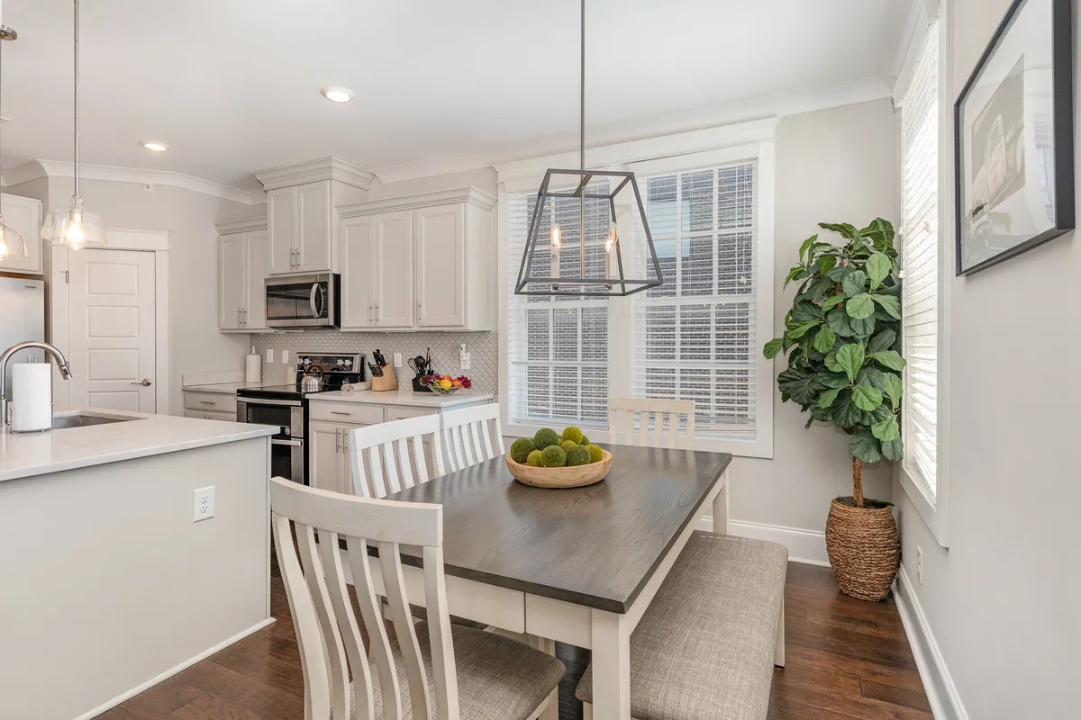 A spacious, sunlit dining area seamlessly connected to a modern kitchen. The dining table is set and ready for a lively meal, with a stylish pendant light overhead and a window letting in natural light. A houseplant in the corner adds a touch of vibrancy to the inviting, communal atmosphere.
