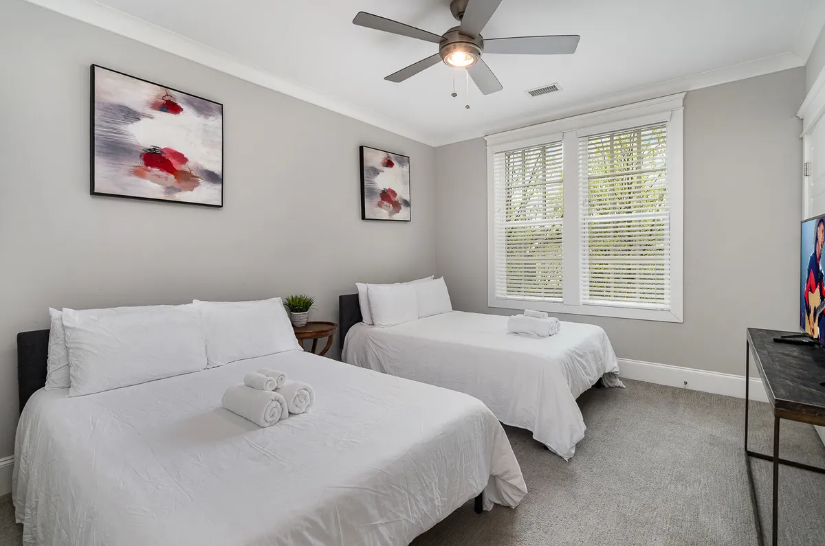 A restful bedroom with two beds dressed in crisp white linens, abstract art adorning the walls, and a window with blinds filtering gentle sunlight. The space feels open and perfect for sharing with family or friends.