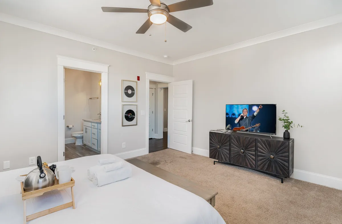 A cozy bedroom with crisp white linens on the bed, a television displaying a musical performance, and a decorative cabinet. The setting feels contemporary and comfortable, inviting guests to relax and enjoy entertainment before sleep.