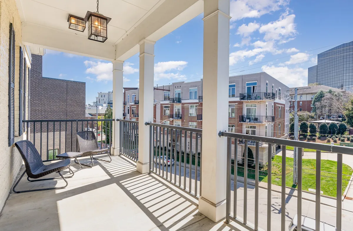 A serene exterior view of a balcony with a comfortable seating area overlooking landscaped greenery. The mood is tranquil, perfect for enjoying morning coffee, reading, or simply relaxing outdoors with a loved one or pet.