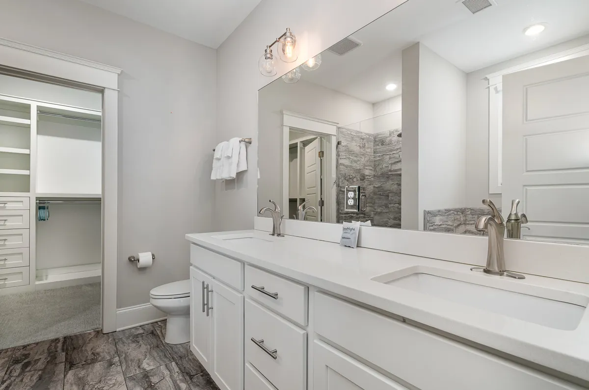 A spacious bathroom featuring a double sink vanity, a large mirror, and an adjacent walk-in closet. The space is organized and serene, designed for both personal grooming and easy organization.